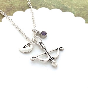 May include: Silver necklace with a bow and arrow charm, a small initial charm, and a purple gemstone.