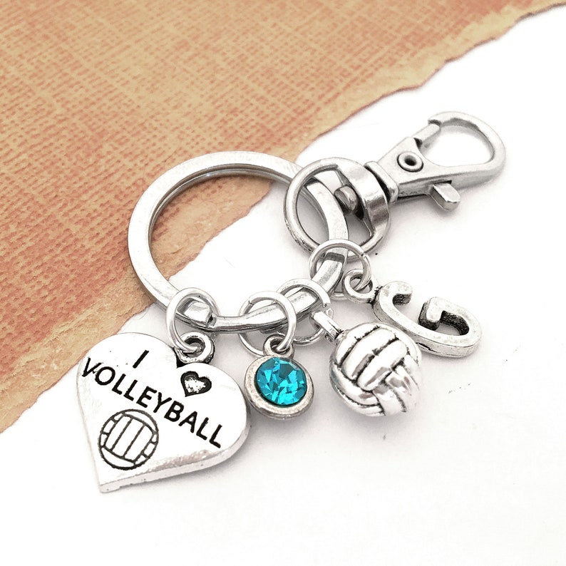 May include: Silver keychain with a heart charm that says "I love volleyball" and a volleyball charm. The keychain also has a blue crystal charm and a silver charm with the letter "G".