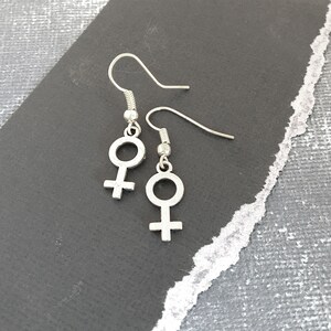 May include: A pair of silver earrings with a female symbol charm. The earrings are on a gray background.