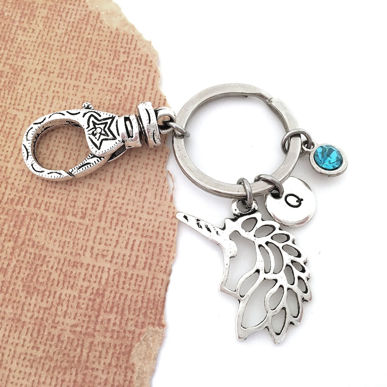 Puede incluir: Llavero de plata con un dije de unicornio, un dije de cristal azul y un dije de plata con la letra "G".