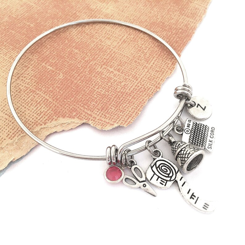 Op de afbeelding: Een zilveren armband met verschillende bedels, waaronder een vingerhoed, een schaar, een spoel garen en een bedel met de tekst "Silk Cord".