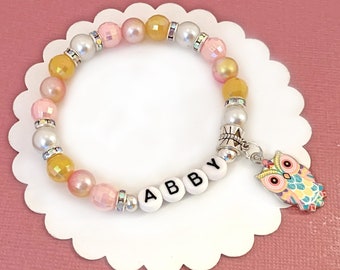 May include: A pink, yellow, and white beaded bracelet with a silver charm that says "ABBY" and a colorful owl charm.