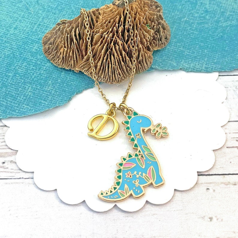 May include: Gold-coloured necklace with a blue dinosaur pendant, decorated with pink and green floral patterns. The necklace also has a gold letter "D" charm. The necklace is presented on a white, scalloped-edge surface.