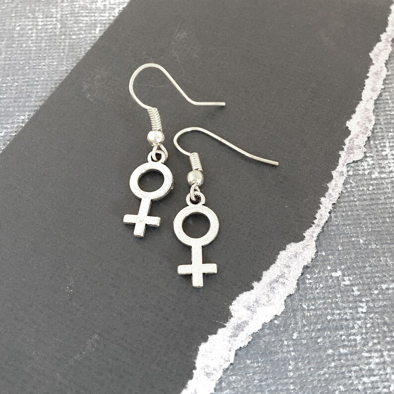 May include: A pair of silver earrings with a female symbol charm. The earrings are on a gray background.