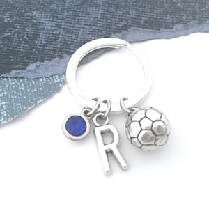 May include: Silver keychain with a blue rhinestone, a silver letter R charm, and a silver soccer ball charm.
