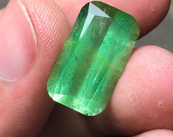 May include: A rectangular, faceted gemstone with a vibrant green hue. The gem has a polished surface with visible internal structures and a brilliant, reflective top facet. The stone is held between fingers, showcasing its size and clarity.