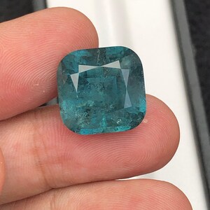May include: A square-cut, teal gemstone with a faceted surface. The gem is held between fingers, showcasing its color and clarity. The stone has a slightly rounded shape and a polished appearance, reflecting light.