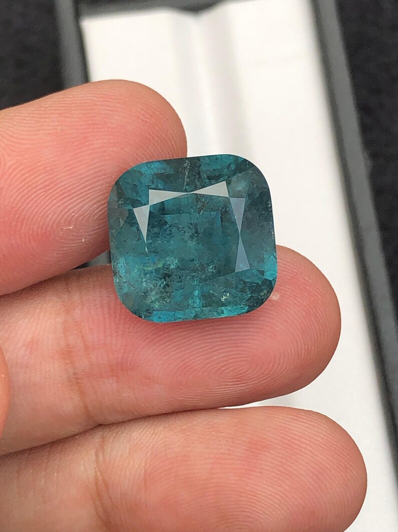 May include: A square-cut, teal gemstone with a faceted surface. The gem is held between fingers, showcasing its color and clarity. The stone has a slightly rounded shape and a polished appearance, reflecting light.
