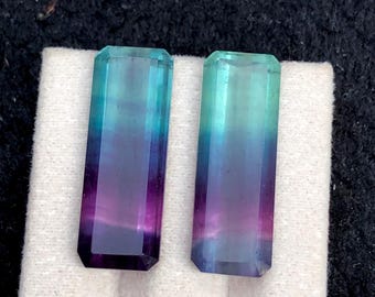 May include: Two rectangular, faceted gemstones with a colour gradient. The stones transition from purple to blue to teal. They are set on a white surface against a dark background. The gemstones are likely for jewellery making.