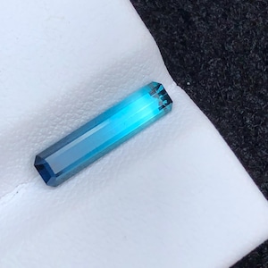Op de afbeelding: Een rechthoekige, gefacetteerde edelsteen met een verloop van blauwtinten, van diep teal tot licht turkoois. De edelsteen wordt gepresenteerd op een wit oppervlak, met een donkergrijze, getextureerde achtergrond.