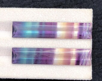 Könnte beinhalten: Zwei rechteckige, mehrfarbige Fluorit-Edelsteine. Die Steine zeigen Bänder in Lila, Blau und Gelb. Die Edelsteine liegen auf einer weißen Oberfläche vor einem dunklen Hintergrund.