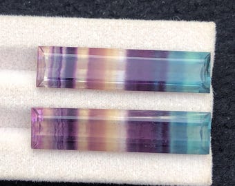 Könnte beinhalten: Zwei rechteckige Fluorite-Edelsteine. Die Edelsteine zeigen Bänder in den Farben Lila, Blau, Gelb und Klar. Die Steine liegen auf einer weißen Oberfläche mit dunklem Hintergrund. Die Edelsteine sind mit geraden Kanten und einer flachen Oberseite geschnitten.