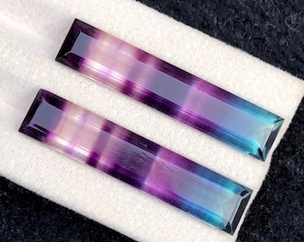 Könnte beinhalten: Zwei rechteckige Fluorite mit einem Farbverlauf aus Lila, Blau und Weiß. Die Steine liegen auf einem weißen Tuch, wodurch ihre polierten Oberflächen und ihre geometrische Form hervorgehoben werden. Ideal für die Schmuckherstellung.