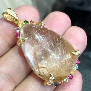 May include: A teardrop-shaped rutilated quartz pendant set in a gold-toned metal frame. The gemstone features visible golden needle-like inclusions. The frame is accented with small, multi-colored round gemstones including red, blue, and green.