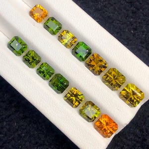 May include: An assortment of square-cut gemstones in shades of green, yellow, and orange are arranged on a white surface. The gemstones are displayed in rows, showcasing their various colors and facets. The gems are likely for jewelry making.