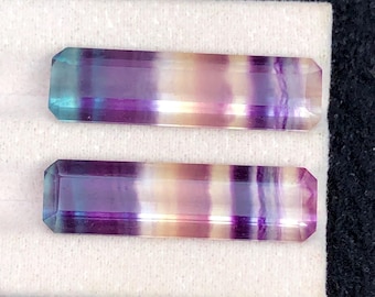 Könnte beinhalten: Zwei rechteckige Fluorite mit violetten, blauen und gelben Farbtönen. Die Edelsteine sind facettiert und poliert. Sie werden auf einer weißen Oberfläche präsentiert.