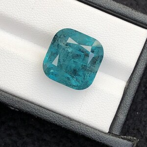 May include: A cushion-cut, teal-colored gemstone displayed in a black box with a white interior. The gemstone has a slightly textured surface and internal reflections, giving it a unique appearance. The box provides a contrasting backdrop, highlighting the gem's color and shape.