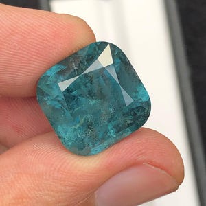 May include: A cushion-cut, teal-colored gemstone with facets that reflect light. The gem is held between fingers, showcasing its clarity and color. The gemstone's shape is a modified square with rounded corners, ideal for jewelry.
