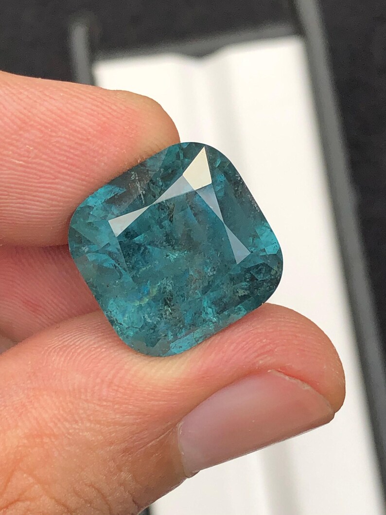 May include: A cushion-cut, teal-colored gemstone with facets that reflect light. The gem is held between fingers, showcasing its clarity and color. The gemstone's shape is a modified square with rounded corners, ideal for jewelry.