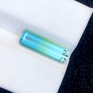 Può includere: Una gemma rettangolare con una sfumatura di colori, che passa dal blu al verde. La gemma è sfaccettata, con molteplici piani che riflettono la luce. Poggia su una superficie bianca, con uno sfondo scuro.