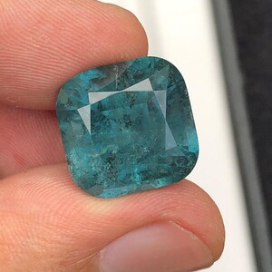 May include: A square-cut, teal-colored gemstone with facets. The gem is held between fingers, showcasing its clarity and color. The stone has a slightly mottled appearance, adding to its unique character.