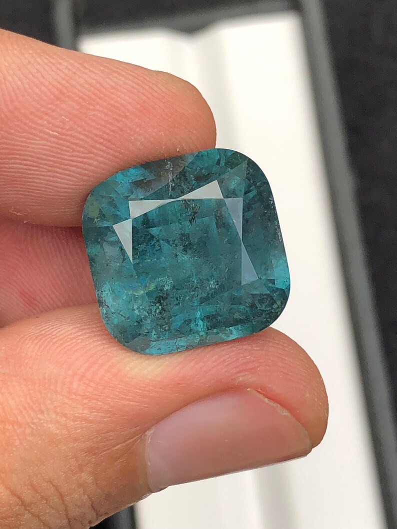 May include: A square-cut, teal-colored gemstone with facets. The gem is held between fingers, showcasing its clarity and color. The stone has a slightly mottled appearance, adding to its unique character.