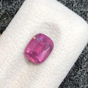 May include: A cushion-cut gemstone with a vibrant, saturated pink hue. The stone features a faceted surface that reflects light, creating distinct geometric patterns across its top. It is positioned on a soft, off-white, textured display pad.