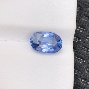 May include: An oval-cut blue sapphire gemstone with a faceted surface that reflects light. The stone displays a vibrant cornflower blue hue and is positioned on a white textured display card, showcasing its clear transparency and polished finish.