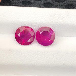 May include: Two round, faceted, vibrant pink gemstones with a brilliant cut. The stones exhibit a deep, saturated magenta hue and are positioned side-by-side, showcasing multiple triangular facets that reflect light across their circular surfaces.