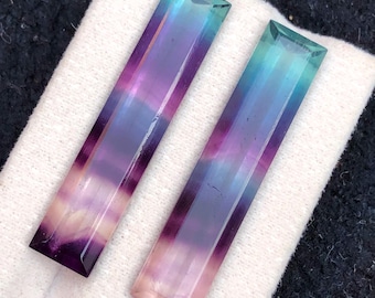 Könnte beinhalten: Zwei rechteckige Fluorite. Die Edelsteine zeigen einen Farbverlauf aus Lila, Blau und Rosa. Die Steine werden auf einem weißen Tuch präsentiert.