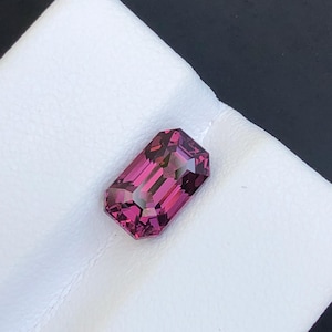 May include: A loose, emerald-cut gemstone with a deep magenta and purple hue. The stone features a faceted surface that reflects light, showcasing a rectangular shape with clipped corners and a transparent, crystalline structure.