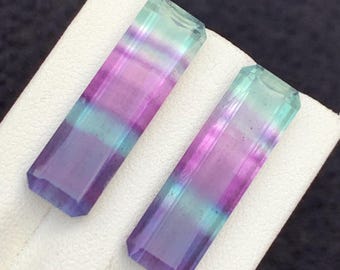 Könnte beinhalten: Zwei rechteckige Fluorite mit Farbverlauf, darunter Lila, Türkis und Klar. Die Edelsteine sind facettiert und haben ein poliertes Aussehen. Sie werden auf einer weißen Oberfläche vor dunklem Hintergrund präsentiert.