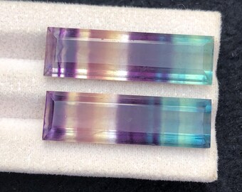 Könnte beinhalten: Zwei rechteckige Fluorite mit Farbverlauf. Die Steine gehen von Lila über Gelb zu Grün und Blau über. Die Edelsteine liegen auf einer weißen Oberfläche.