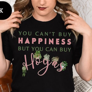 May include: A black t-shirt with the text "You can't buy happiness but you can buy Hoyas" in pink lettering. The shirt features a graphic of several potted hoya plants.