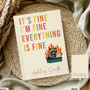 May include: Ivory-colored notebook with the text "IT'S FINE I'M FINE EVERYTHING IS FINE" in colorful letters. A raccoon with a laptop sits in front of flames. The name "Ashley Smith" is written below.