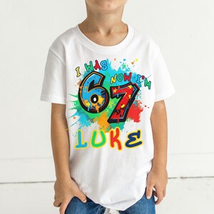 May include: White t-shirt with a colorful graphic design. The design includes the text "I WAS 6 NOW I'M 7 LUKE" in various colors, with paint splatter accents. The shirt is worn by a child.