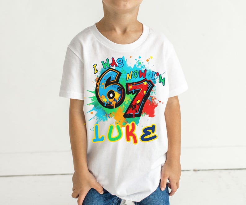 May include: White t-shirt with a colorful graphic design. The design includes the text "I WAS 6 NOW I'M 7 LUKE" in various colors, with paint splatter accents. The shirt is worn by a child.