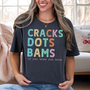 May include: A dark gray t-shirt with the words "CRACKS DOTS BAMS" in colorful block letters. Below the words is the phrase "If you know you know" in a smaller font. The shirt is worn by a person.