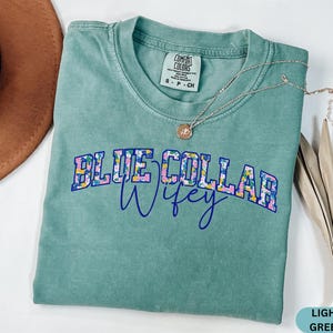 May include: A light green t-shirt with the words "BLUE COLLAR Wifey" in a floral design. A silver necklace with a pendant is draped across the shirt. A brown hat is partially visible in the background.