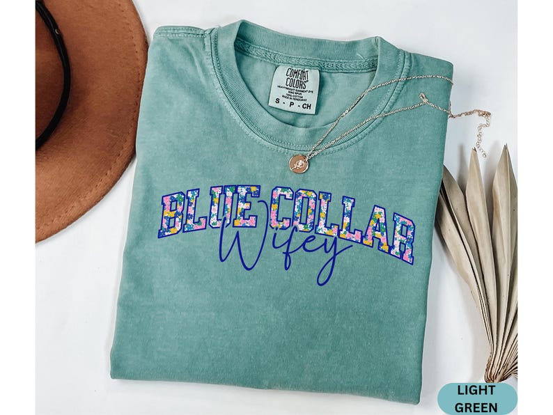 May include: A light green t-shirt with the words "BLUE COLLAR Wifey" in a floral design. A silver necklace with a pendant is draped across the shirt. A brown hat is partially visible in the background.