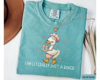 May include: Seafoam green Comfort Colors t-shirt featuring a cartoon goose wearing a chef's hat and apron, carrying cupcakes. The text "I AM LITERALLY JUST A BAKER" is printed below the goose. A gold necklace is draped across the shirt.