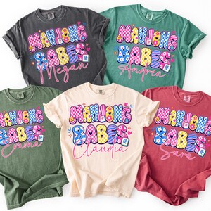 May include: Five colorful t-shirts with the text "Mahjong Babes" in a playful, bubble font. Each shirt features a different name in a cursive font. The shirts are in various colors, including charcoal, green, cream, red, and teal.