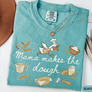 May include: Seafoam green t-shirt with the phrase "Mama makes the dough" and a cartoon duck wearing a chef's hat and apron, carrying a loaf of bread. The shirt features baking-related illustrations.
