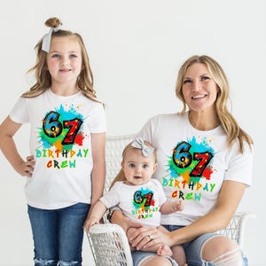 May include: Three people wearing matching white t-shirts with a colorful "67 BIRTHDAY CREW" design. The shirts have a paint splatter effect. The people are a young girl, a baby, and an adult woman. The girl and woman are wearing blue jeans.