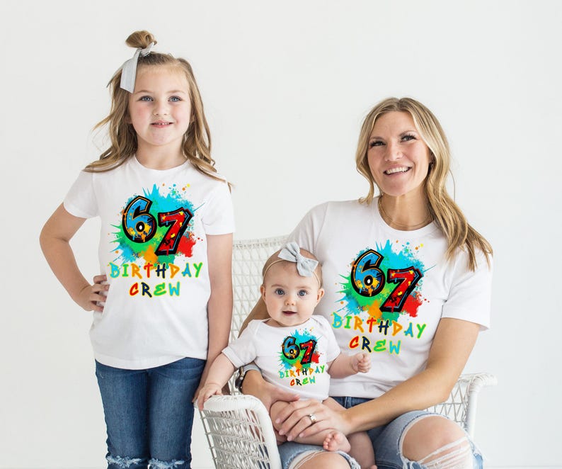 May include: Three people wearing matching white t-shirts with a colorful "67 BIRTHDAY CREW" design. The shirts have a paint splatter effect. The people are a young girl, a baby, and an adult woman. The girl and woman are wearing blue jeans.