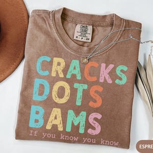 May include: Espresso-colored t-shirt with the words "CRACKS DOTS BAMS" in colorful block letters. The text "If you know you know" is below the main text. A silver necklace and a brown hat are also visible.