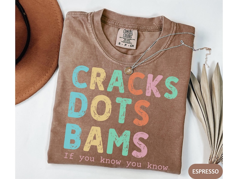 May include: Espresso-colored t-shirt with the words "CRACKS DOTS BAMS" in colorful block letters. The text "If you know you know" is below the main text. A silver necklace and a brown hat are also visible.