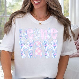 K&ouml;nnte beinhalten: Wei&szlig;es T-Shirt mit dem Text "Weenie Mom" in einem Blumen- und Vichy-Muster. Eine Dackel-Grafik befindet sich in der Mitte. Es ist ein l&auml;ssiges Kleidungsst&uuml;ck f&uuml;r den Alltag.