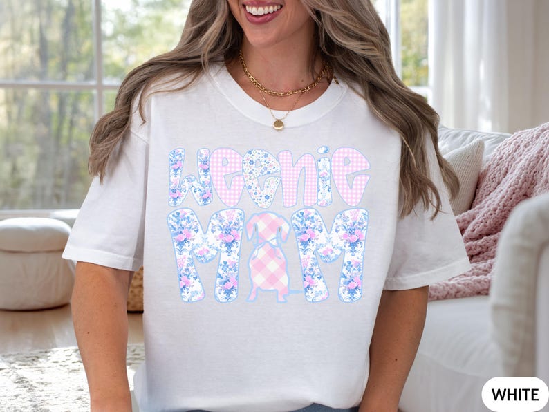 K&ouml;nnte beinhalten: Wei&szlig;es T-Shirt mit dem Text "Weenie Mom" in einem Blumen- und Vichy-Muster. Eine Dackel-Grafik befindet sich in der Mitte. Es ist ein l&auml;ssiges Kleidungsst&uuml;ck f&uuml;r den Alltag.