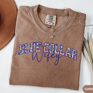 May include: A brown t-shirt with the text "BLUE COLLAR Wifey" in a floral design. A silver necklace with a pendant is on the shirt. A brown hat is visible in the upper left corner. The shirt is a Comfort Colors brand.